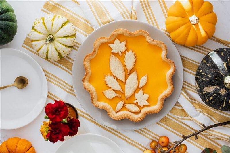 A pumpkin pie with a golden filling and leaf-shaped pastry decorations on a plate, surrounded by small pumpkins and flowers