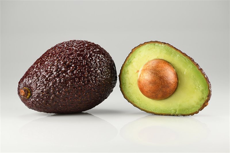 A whole dark Hass avocado beside a halved avocado showing its large round pit